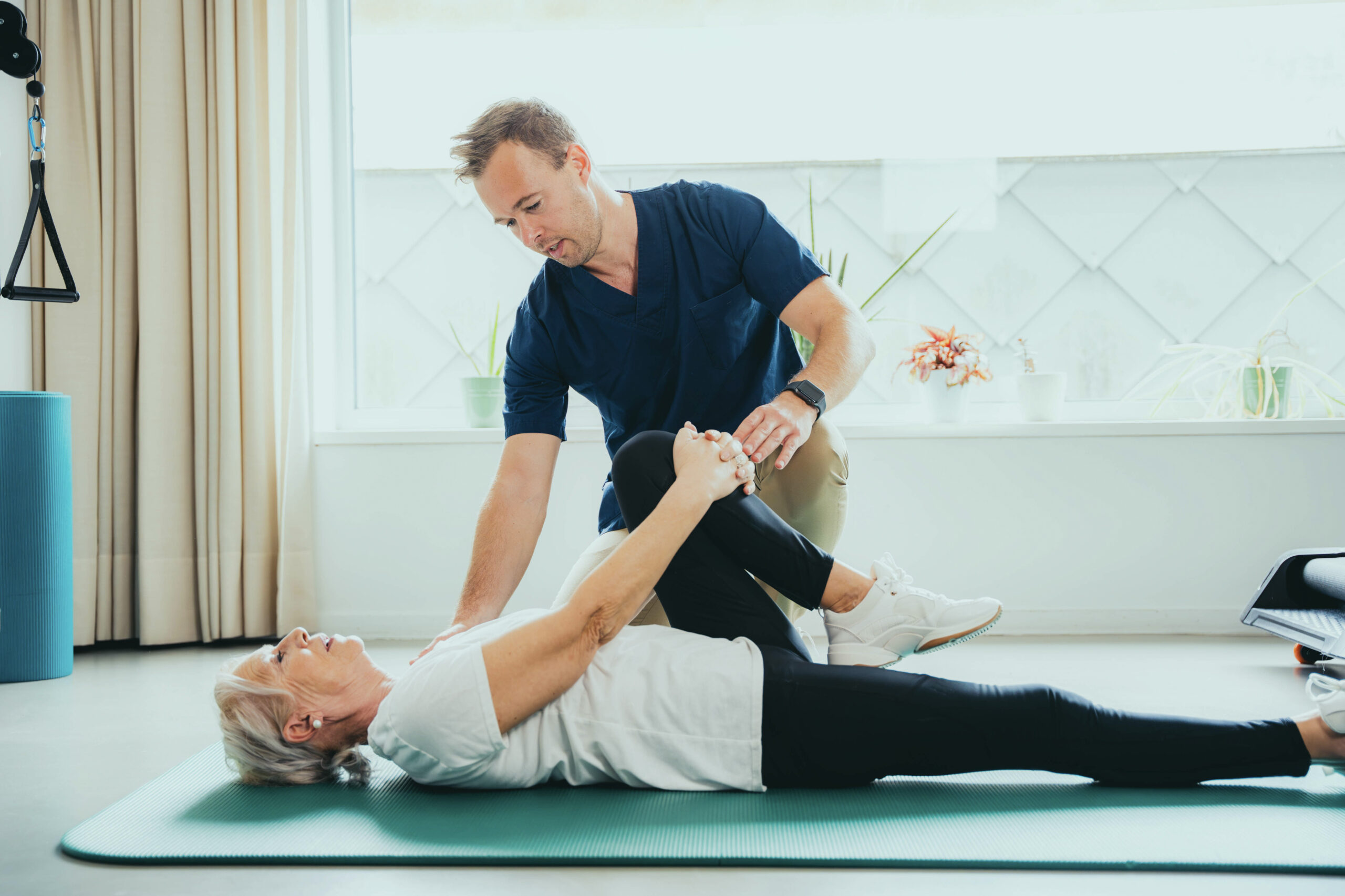 Physical therapist guiding an older adult through a gentle knee to chest stretch during hip rehab on a clinic mat
