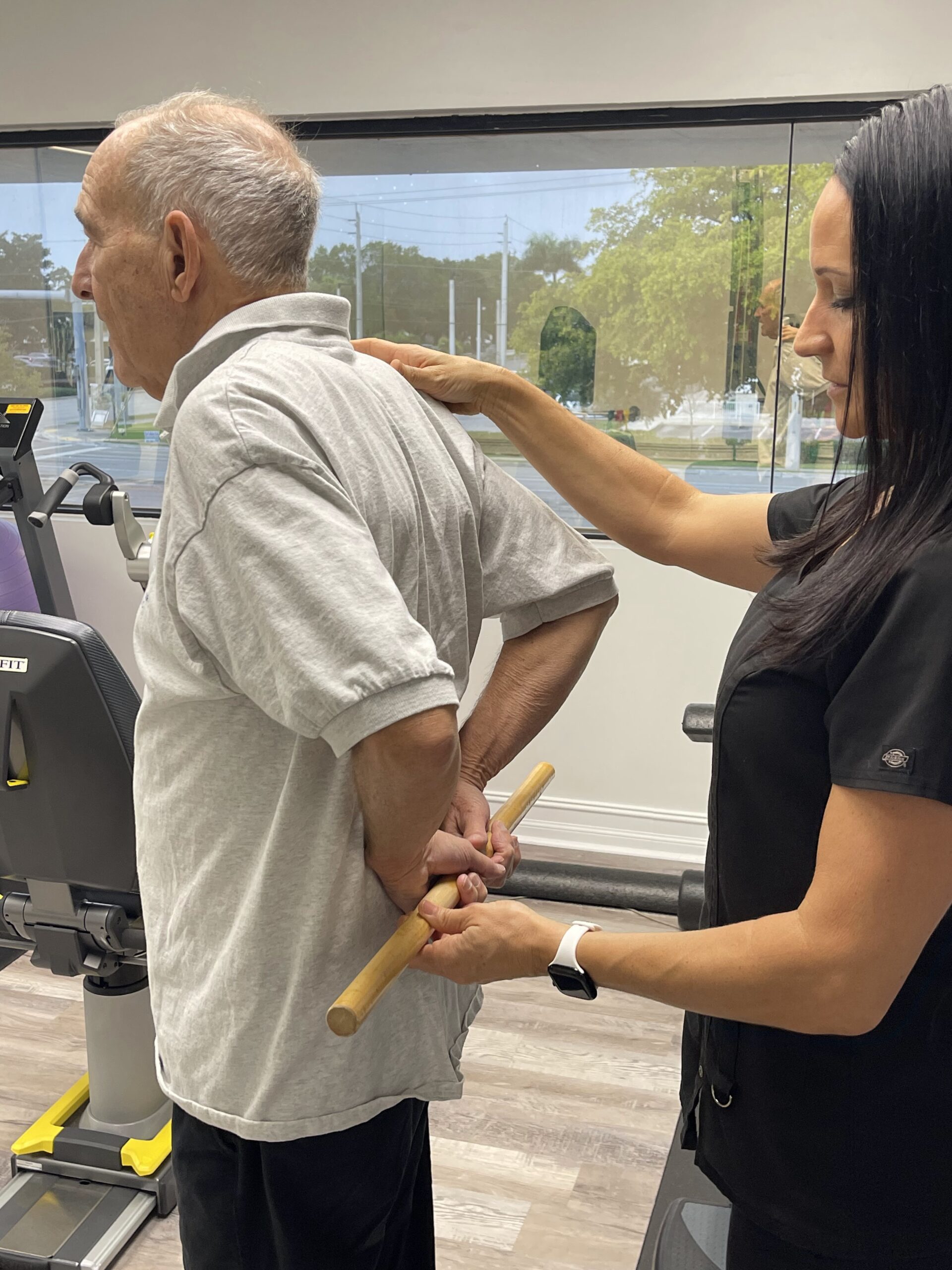 Senior man receiving guided physical therapy support while using a wooden dowel to improve shoulder mobility at a rehab center.