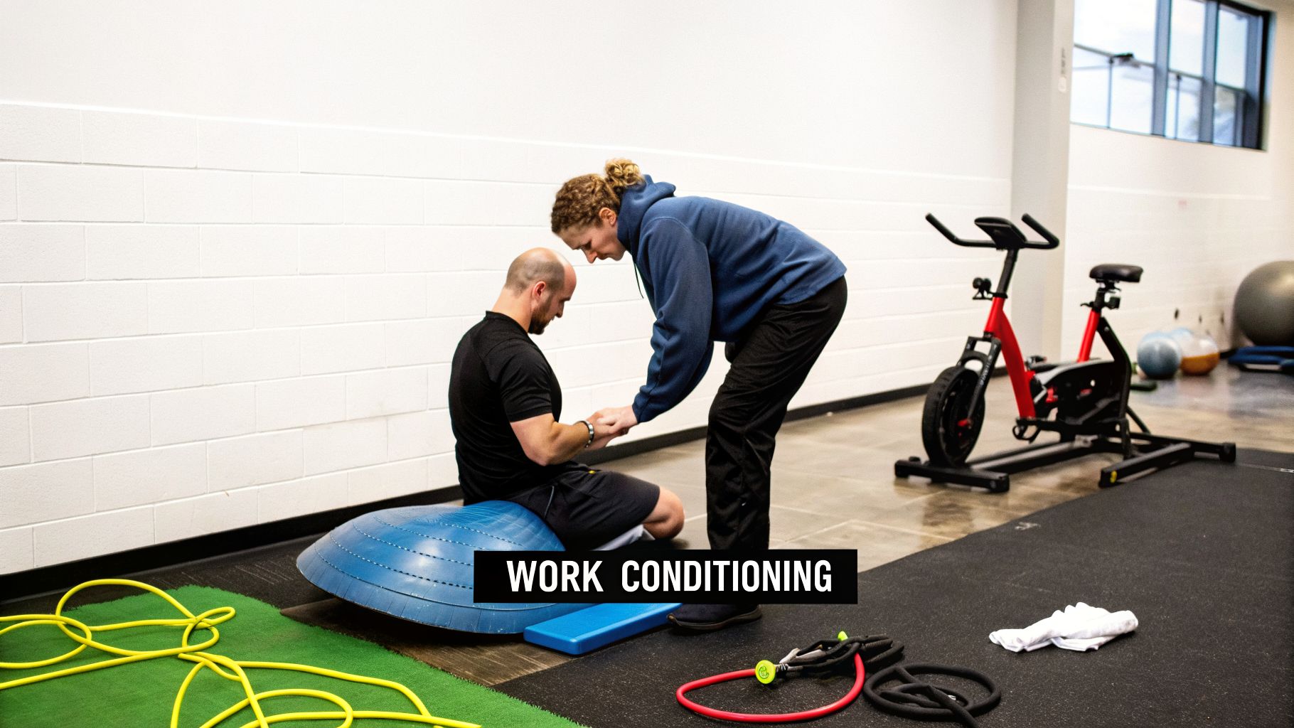 A man sits on a BOSU ball, guided by a woman, during a work conditioning rehabilitation session in a gym.