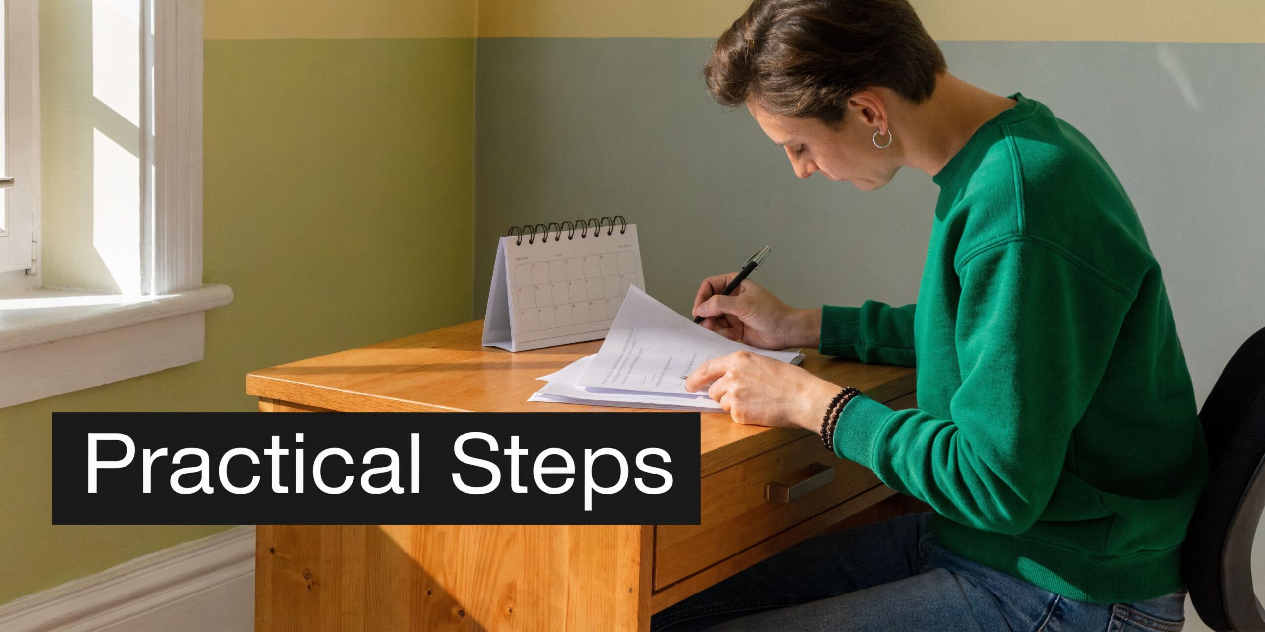 A person wearing a green sweater sits at a wooden desk writing on documents near a calendar.