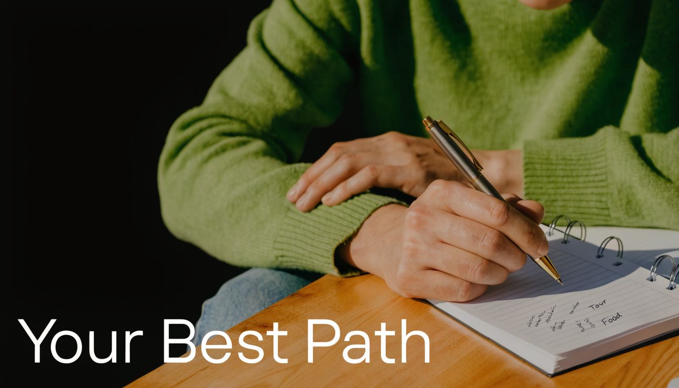 A person wearing a green sweater writes in a notebook with a pen at a wooden desk.