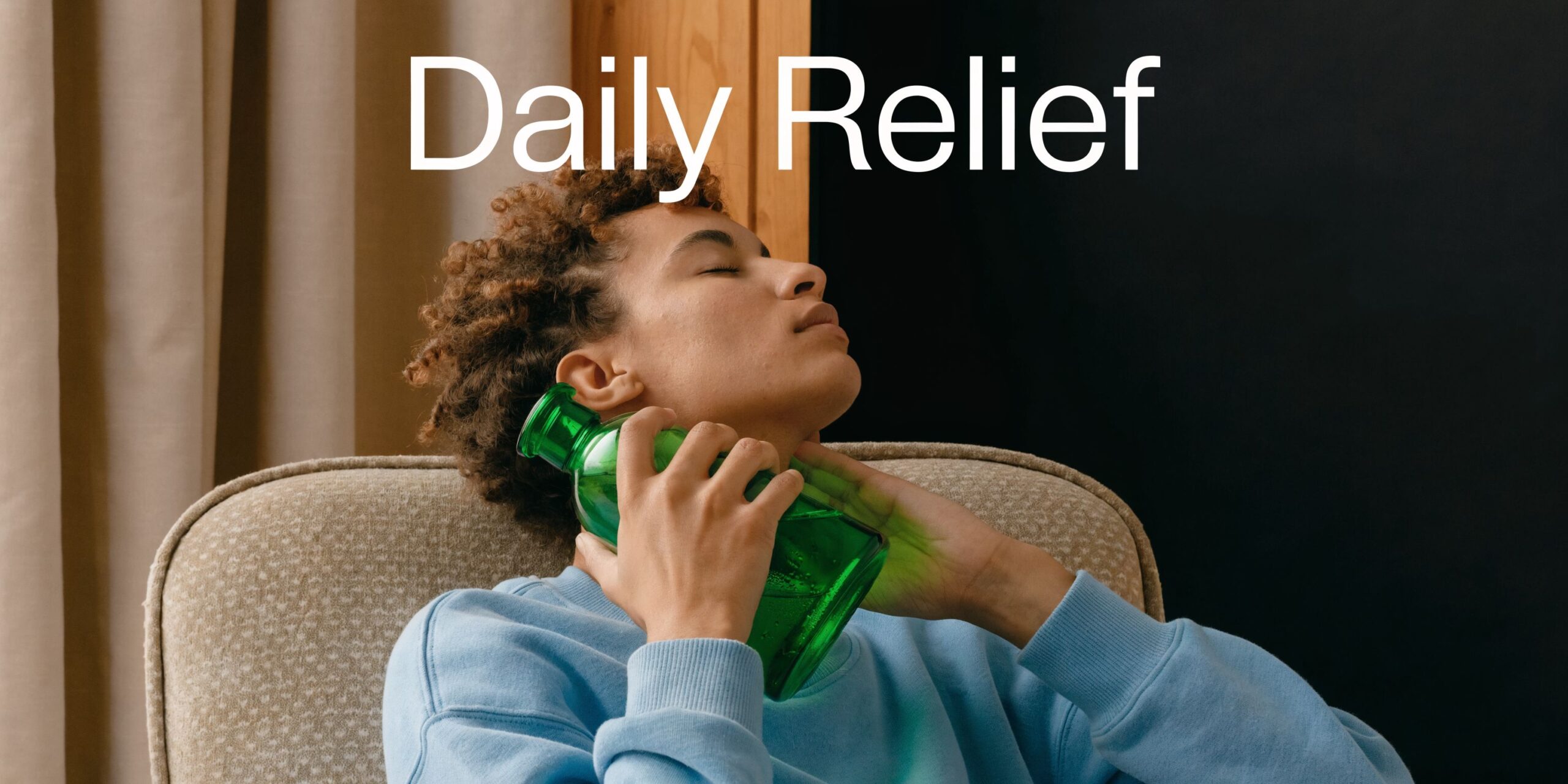 A person with curly hair relaxing in a chair while holding a green bottle against their neck.