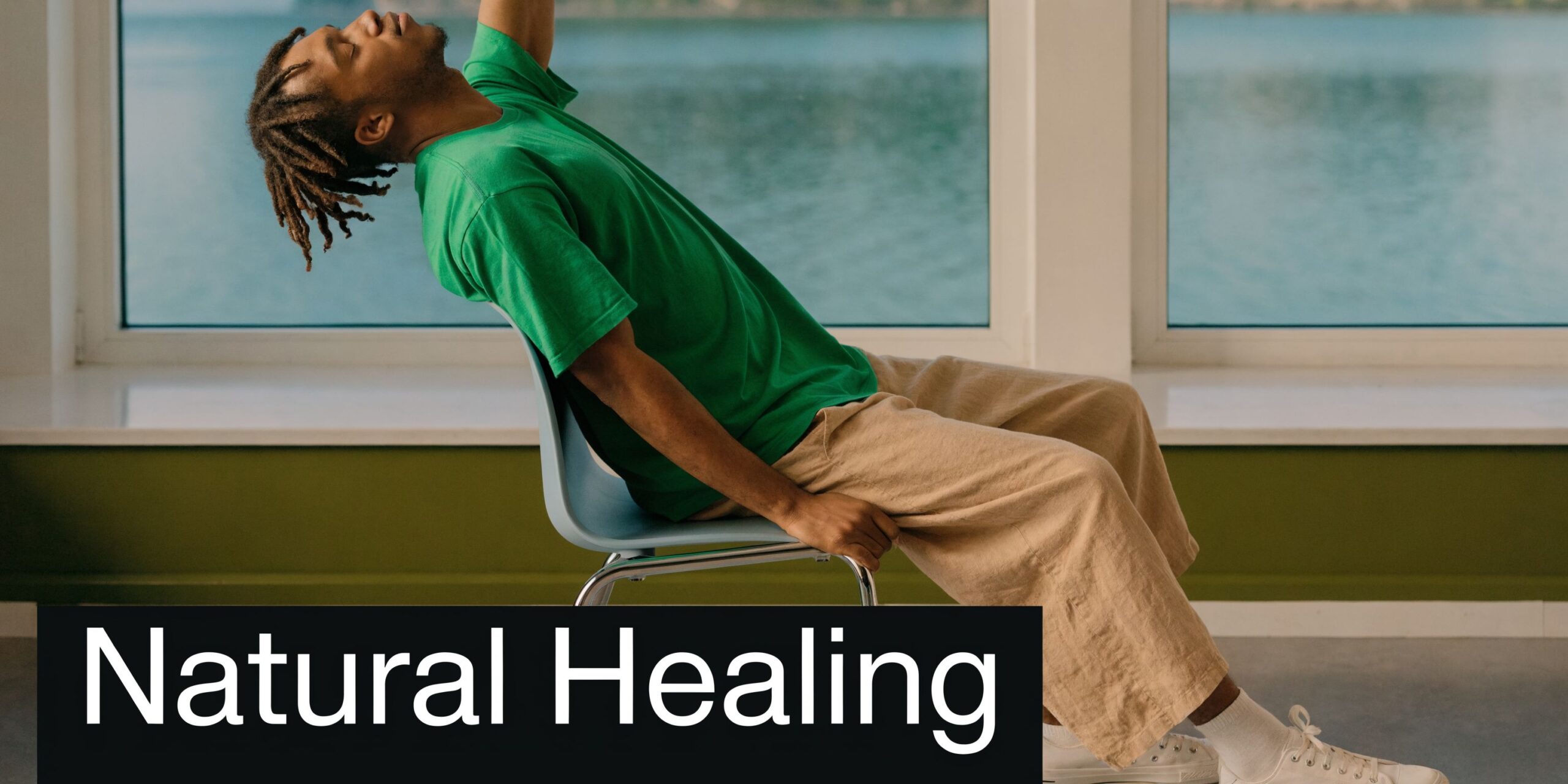 A young man with dreadlocks wearing a green t-shirt stretches his back while sitting on a chair.