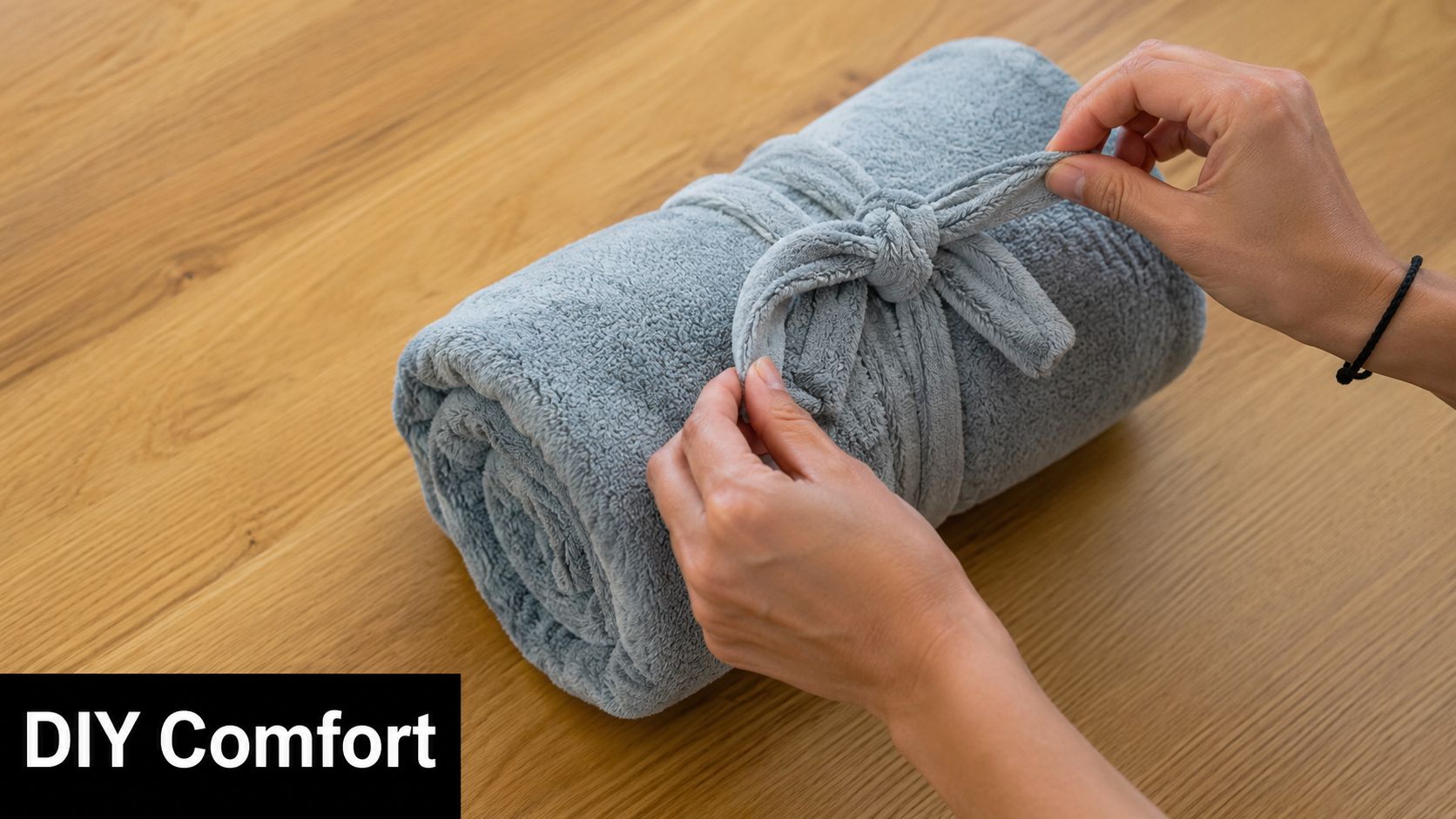 A person wrapping a soft blue fleece blanket into a neat roll on a wooden table.